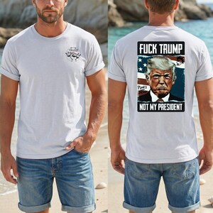 Op de afbeelding: Lichtgrijs t-shirt met opgerolde mouwen. De voorkant heeft een kleine afbeelding en tekst. De achterkant toont een afbeelding van Donald Trump met de tekst "FUCK TRUMP NOT MY PRESIDENT" in een zwarte rechthoek met een vlagachtergrond.