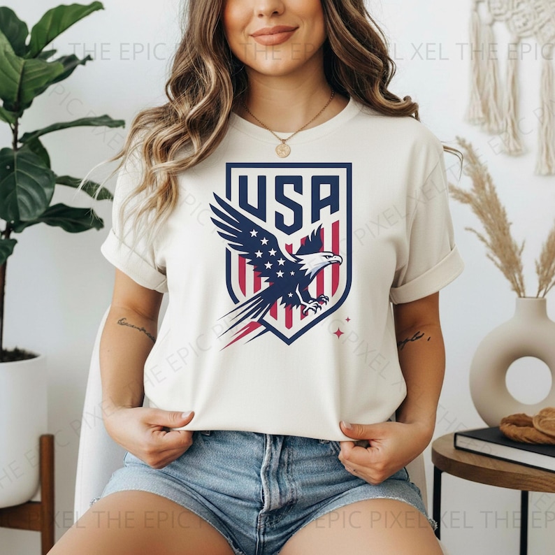 May include: Cream-colored t-shirt featuring a shield design with the text "USA" in navy blue. The shield showcases a bald eagle with a red, white, and blue color scheme. The shirt is a short-sleeved crew neck.