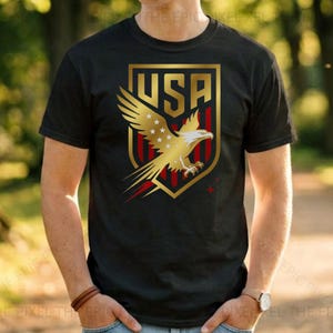 May include: Black t-shirt featuring a gold and red USA emblem with a golden eagle design. The emblem includes the word "USA" in gold lettering. The shirt is a crew neck style.