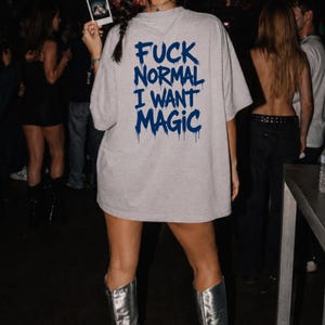 Op de afbeelding: Een lichtgrijs T-shirt met korte mouwen en de donkerblauwe tekst "FUCK NORMAL I WANT MAGIC" op de achterkant in een gestileerd, druipend lettertype. Het T-shirt heeft een losse, oversized pasvorm en wordt gedragen met zilveren laarzen.