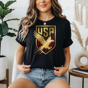May include: Black t-shirt featuring a gold and red USA emblem with an eagle design. The emblem is set within a gold shield shape. The shirt is a crew neck style.