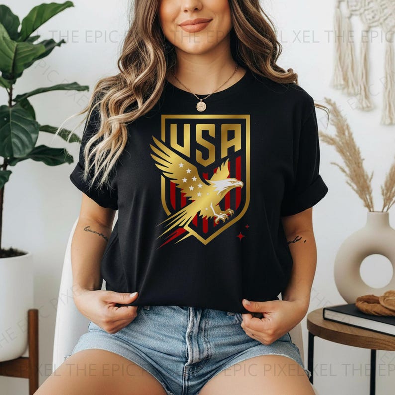 May include: Black t-shirt featuring a gold and red USA emblem with an eagle design. The emblem is set within a gold shield shape. The shirt is a crew neck style.