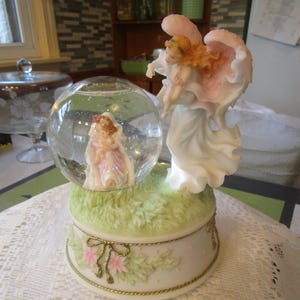 May include: A decorative angel figurine with a snow globe. The snow globe features a small angel figure inside. The base is decorated with pink flowers, green grass, and a gold-colored rope trim. The angel figurine has a white dress and pink wings.