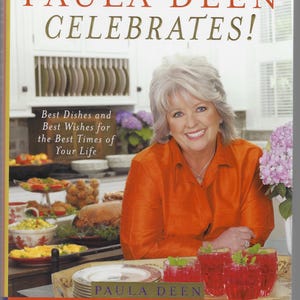 May include: A cookbook cover featuring Paula Deen smiling, wearing an orange shirt. The title reads "Paula Deen Celebrates!" with the subtitle "Best Dishes and Best Wishes for the Best Times of Your Life." Several dishes and drinks are on the table.