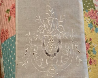 Puede incluir: Un tejido rectangular de lino beige claro con intrincados bordados florales blancos y detalles de calado. El diseño incluye un motivo central de rosa rodeado de patrones de volutas y un borde festoneado con delicada costura.