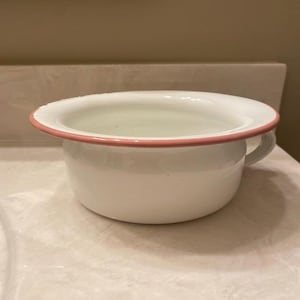 May include: A white enamel chamber pot with a smooth, glossy finish and a distinct pink rim. The basin features a wide, rounded opening and a small, curved handle on the side, presenting a simple, functional design with clean, rounded edges.