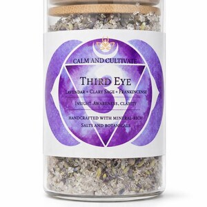 May include: A glass jar filled with bath salts, featuring a wooden lid and a label with the words "Third Eye." The label includes the ingredients: lavender, clary sage, and frankincense. The text "Calm and Cultivate" is also visible.