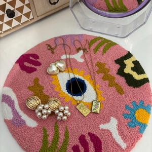 Op de afbeelding: Een roze en wit tapijt met kleurrijke abstracte vormen en een paar gouden oorbellen met parel accenten. Er zijn drie gouden kettingen met hangers, waaronder een hart, een ster en een rechthoek met een ontwerp.