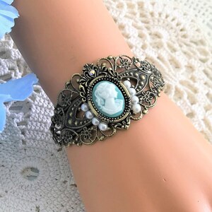 May include: A delicate bracelet featuring a cameo with a woman's profile in a light blue and white design. The bracelet is made of gold-toned metal and features a filigree design with white pearls.