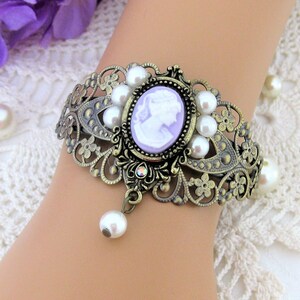 May include: An antique-style bracelet with a cameo design. The bronze-colored metal bracelet features a central oval cameo in lavender, surrounded by faux pearls. A dangling pearl hangs from the bottom, adding a touch of elegance.
