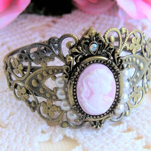 May include: A delicate, ornate, gold-toned metal bracelet with a pink cameo featuring a woman's profile. The bracelet is adorned with small white pearls.