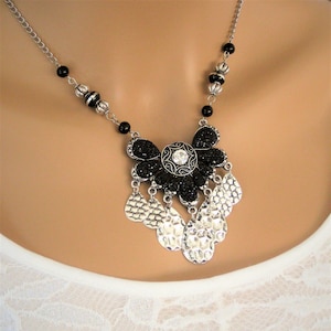 May include: A silver chain necklace with black beads and a large silver pendant featuring a black flower with a rhinestone center and dangling silver teardrop shapes.