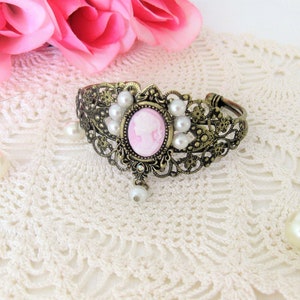 May include: An antique bronze cuff bracelet with a pink cameo center and faux pearls. The bracelet features an oval cameo with a woman's profile, surrounded by small pearls and ornate filigree detailing. A single pearl dangles below the cameo.