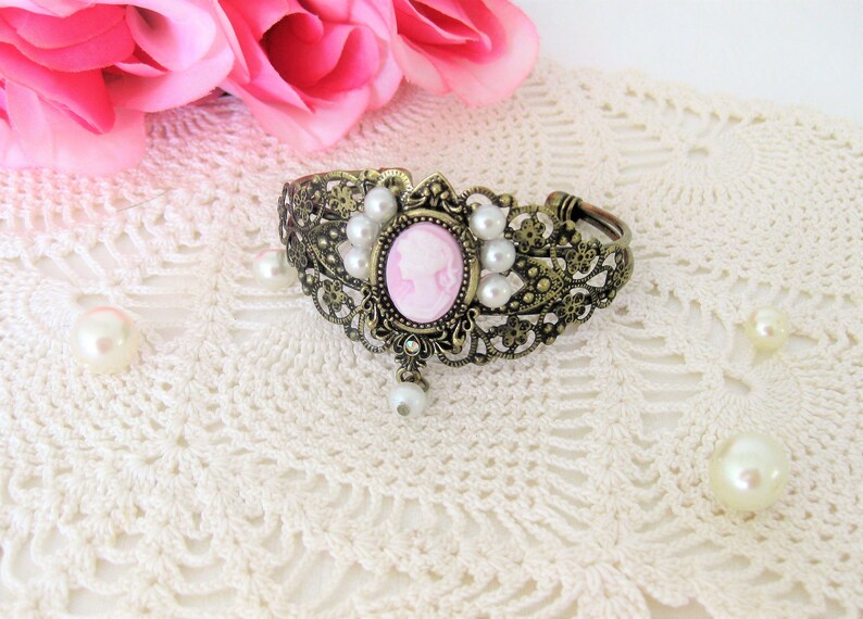 May include: An antique bronze cuff bracelet with a pink cameo center and faux pearls. The bracelet features an oval cameo with a woman's profile, surrounded by small pearls and ornate filigree detailing. A single pearl dangles below the cameo.