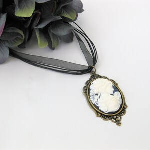 May include: A vintage-style cameo pendant necklace. The oval pendant features a white relief of a woman's profile against a dark blue background, framed by an ornate bronze-colored setting. The necklace has a black ribbon.