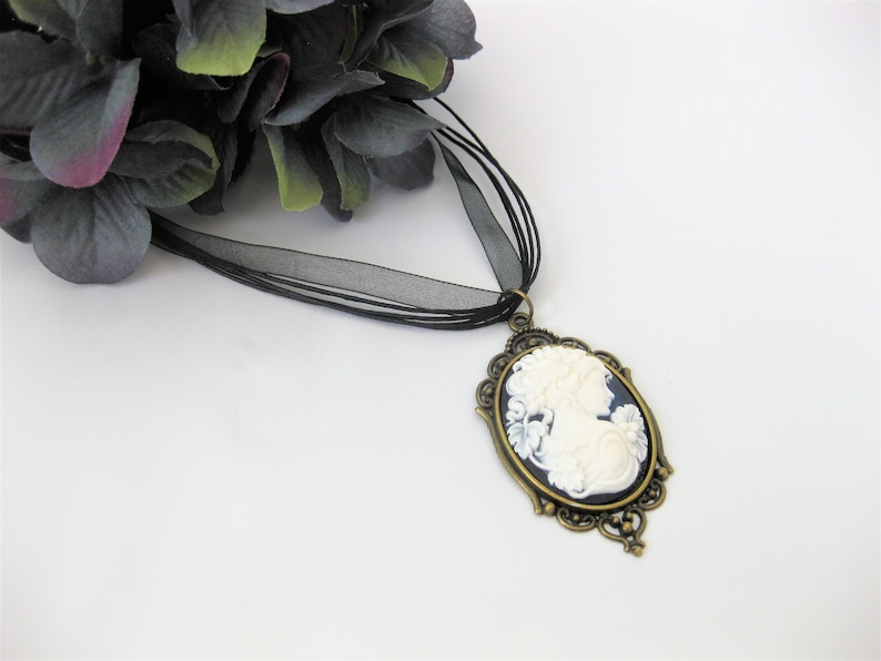 May include: A vintage-style cameo pendant necklace. The oval pendant features a white relief of a woman's profile against a dark blue background, framed by an ornate bronze-colored setting. The necklace has a black ribbon.