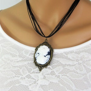 May include: A cameo pendant necklace featuring a white profile against a dark blue background, set in an ornate bronze-toned frame. The necklace hangs from a black ribbon. The pendant is a classic piece of jewelry.