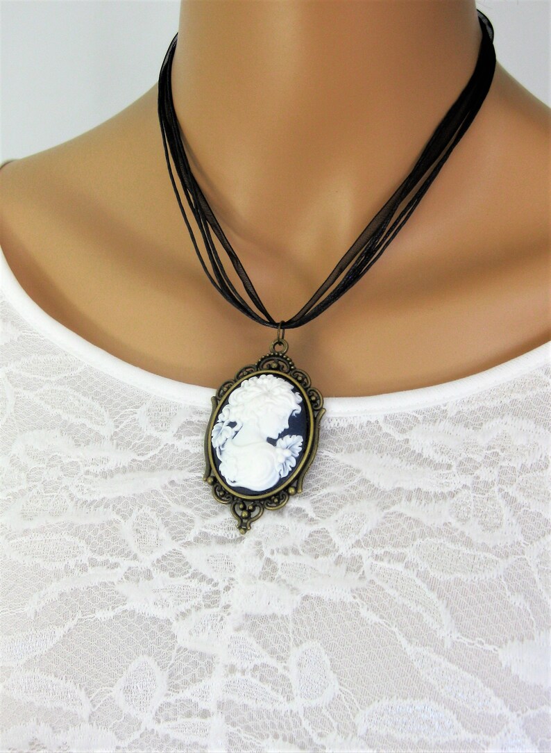 May include: A cameo pendant necklace featuring a white profile against a dark blue background, set in an ornate bronze-toned frame. The necklace hangs from a black ribbon. The pendant is a classic piece of jewelry.