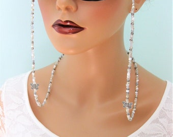 May include: Eyeglass chain with clear and silver beads and two silver butterfly charms. The chain is attached to black framed eyeglasses. The model is wearing a white lace top.