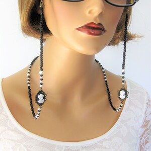 May include: Black and white beaded eyeglass chain with cameo charms. The chain is worn by a model with short brown hair and black framed glasses.