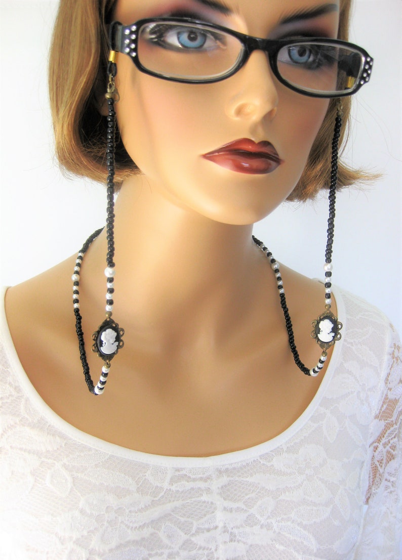 May include: Black and white beaded eyeglass chain with cameo charms. The chain is worn by a model with short brown hair and black framed glasses.