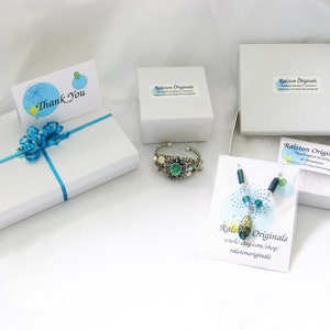 May include: A white gift box with a blue ribbon and a thank you card. The box is surrounded by other white boxes containing jewellery. The jewellery includes a green and silver bracelet and a blue and green necklace. The boxes have the text "Ralston Originals" printed on them.