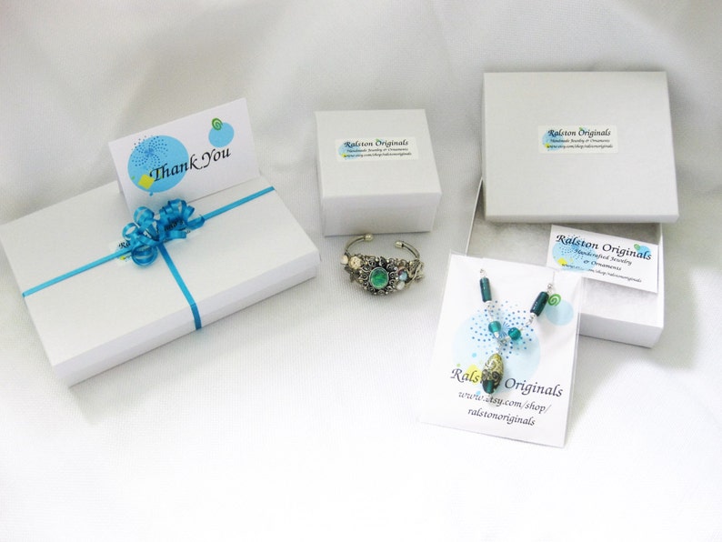 May include: A white gift box with a blue ribbon and a thank you card. The box is surrounded by other white boxes containing jewellery. The jewellery includes a green and silver bracelet and a blue and green necklace. The boxes have the text "Ralston Originals" printed on them.
