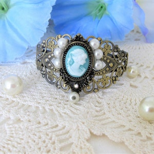 May include: An antique-style bracelet with a cameo design. The bracelet features a central oval cameo with a light blue background and a white profile. It is surrounded by faux pearls and ornate metalwork. The bracelet is set against a white lace background.