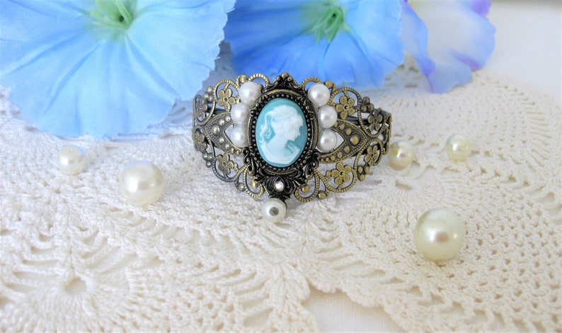May include: An antique-style bracelet with a cameo design. The bracelet features a central oval cameo with a light blue background and a white profile. It is surrounded by faux pearls and ornate metalwork. The bracelet is set against a white lace background.