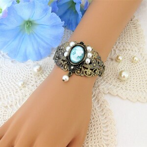 May include: An antique-style bracelet with a cameo design. The bracelet features a bronze-colored filigree design, adorned with faux pearls and a central oval cameo with a light blue profile. A single pearl dangles from the bottom.