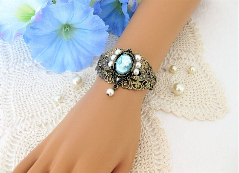 May include: An antique-style bracelet with a cameo design. The bracelet features a bronze-colored filigree design, adorned with faux pearls and a central oval cameo with a light blue profile. A single pearl dangles from the bottom.