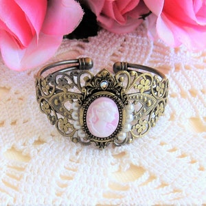 May include: A gold-tone metal bracelet with a delicate floral design. The bracelet features a pink cameo with a woman's profile and is accented with white pearls.