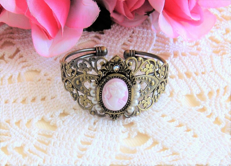 May include: A gold-tone metal bracelet with a delicate floral design. The bracelet features a pink cameo with a woman's profile and is accented with white pearls.