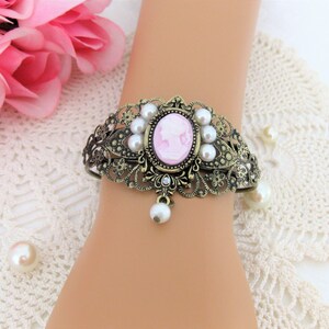 May include: Ornate bronze-toned cuff bracelet featuring a pink cameo with a white silhouette. The bracelet is adorned with faux pearls and a dangling pearl pendant. The design includes intricate filigree and floral details, creating a vintage aesthetic.