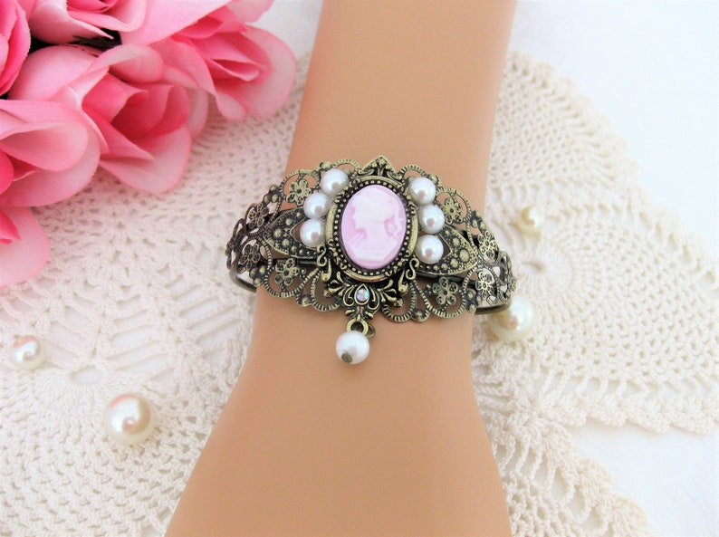 May include: Ornate bronze-toned cuff bracelet featuring a pink cameo with a white silhouette. The bracelet is adorned with faux pearls and a dangling pearl pendant. The design includes intricate filigree and floral details, creating a vintage aesthetic.