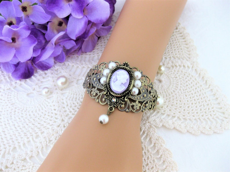 May include: An antique-style bracelet with a cameo design. The bronze-colored metal bracelet features a central oval cameo in lavender, surrounded by faux pearls. A dangling pearl hangs from the bottom, adding a touch of elegance.