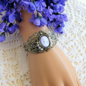 May include: A delicate bracelet featuring a cameo with a woman's profile in a lavender colour. The bracelet is made of gold-toned metal and features a floral design with pearl accents.