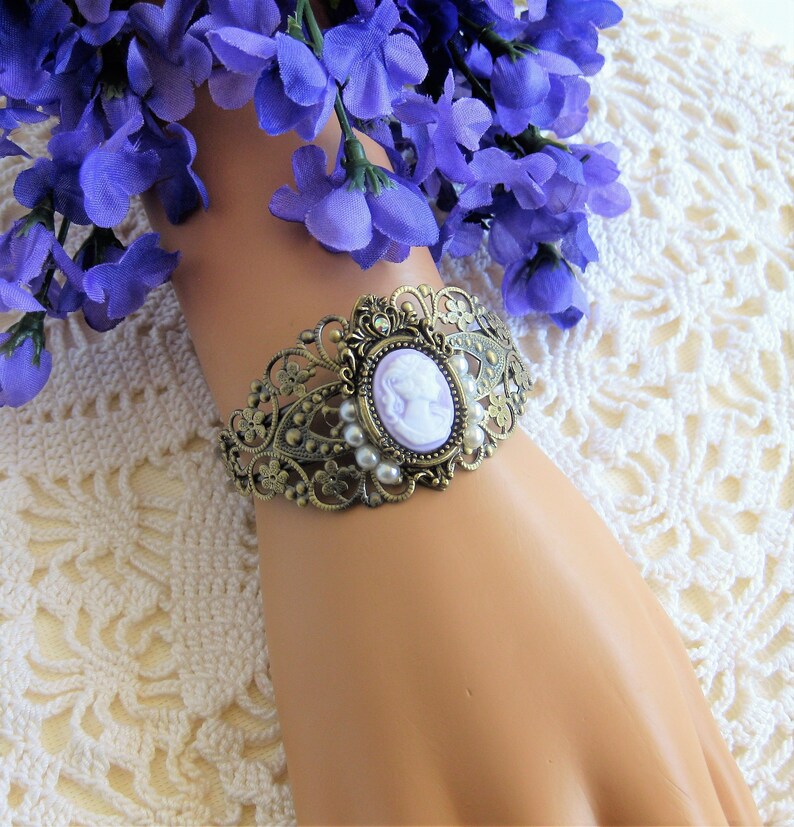 May include: A delicate bracelet featuring a cameo with a woman's profile in a lavender colour. The bracelet is made of gold-toned metal and features a floral design with pearl accents.