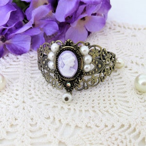 May include: An antique-style bronze bracelet with a cameo in the center, surrounded by faux pearls. The cameo features a white profile on a lavender background. The bracelet has an ornate, filigree design and a dangling pearl charm.