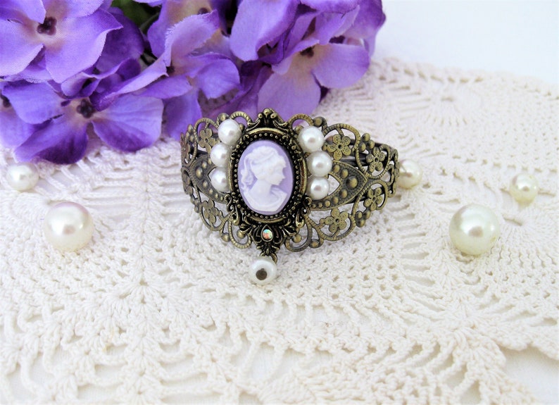 May include: An antique-style bronze bracelet with a cameo in the center, surrounded by faux pearls. The cameo features a white profile on a lavender background. The bracelet has an ornate, filigree design and a dangling pearl charm.