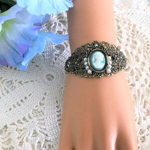 May include: A delicate bracelet featuring a cameo with a woman's profile in white against a light blue background. The bracelet is made of gold-toned metal and features a filigree design with white pearl accents.