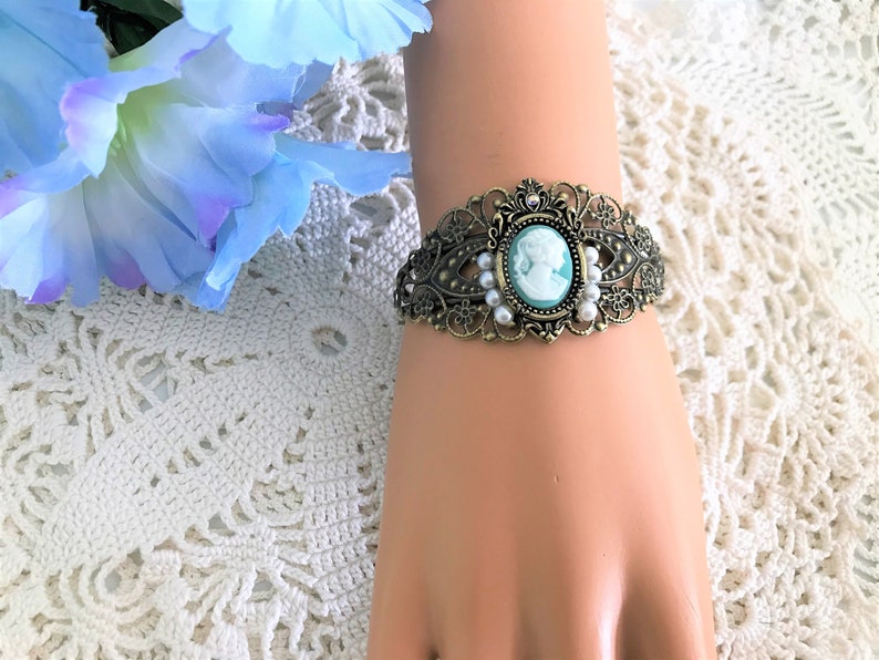 May include: A delicate bracelet featuring a cameo with a woman's profile in white against a light blue background. The bracelet is made of gold-toned metal and features a filigree design with white pearl accents.