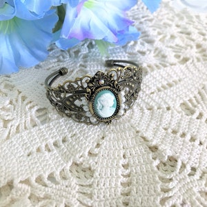 May include: A delicate bronze bracelet with a cameo featuring a woman's profile in white against a light blue background. The bracelet is adorned with small white pearls and a crystal accent.