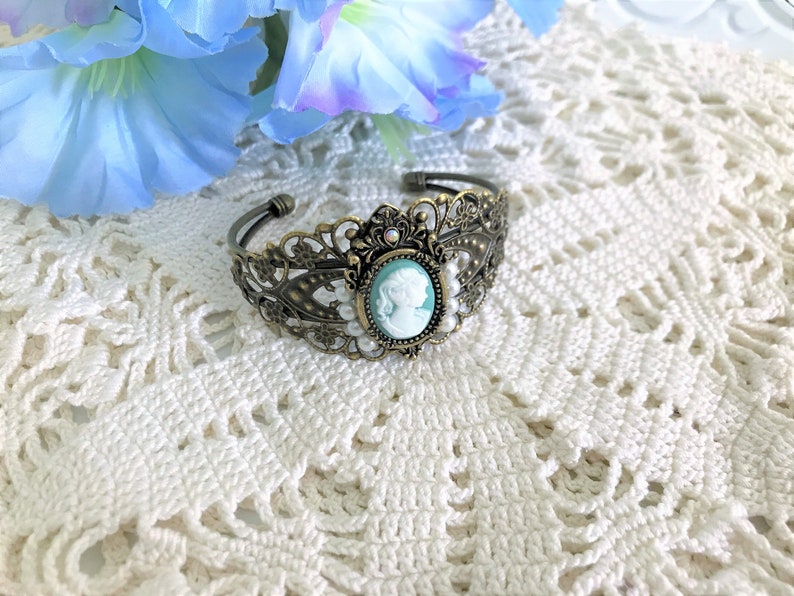 May include: A delicate bronze bracelet with a cameo featuring a woman's profile in white against a light blue background. The bracelet is adorned with small white pearls and a crystal accent.