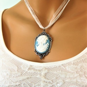 May include: A cameo pendant necklace featuring a white profile on a light blue background, set in an ornate silver-toned frame. The pendant hangs from a sheer white ribbon necklace, creating a classic and elegant look. The necklace is worn over a white lace top.