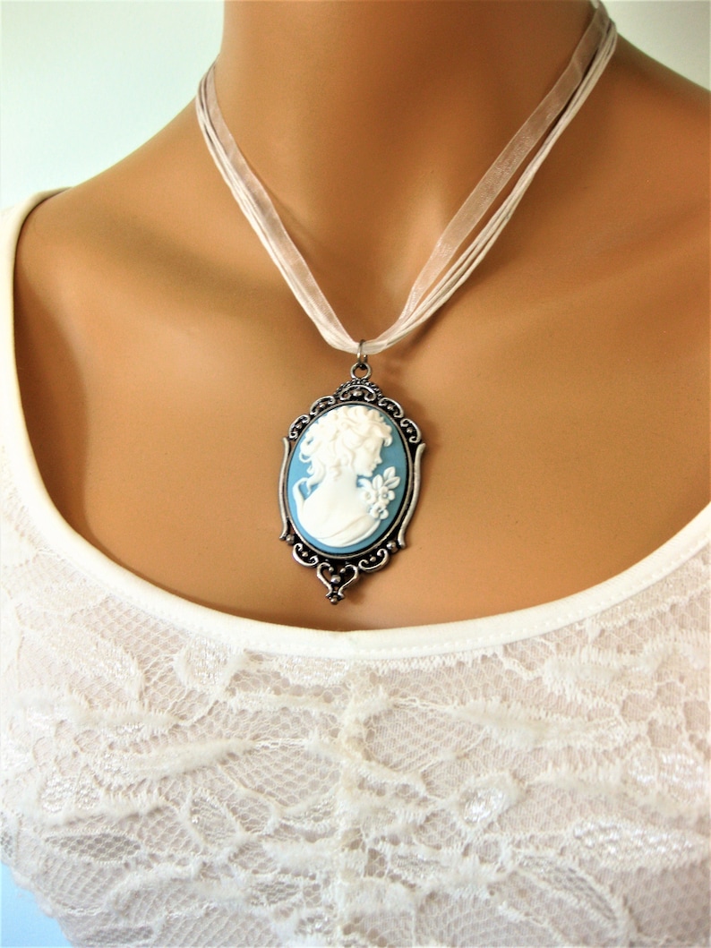 May include: A cameo pendant necklace featuring a white profile on a light blue background, set in an ornate silver-toned frame. The pendant hangs from a sheer white ribbon necklace, creating a classic and elegant look. The necklace is worn over a white lace top.