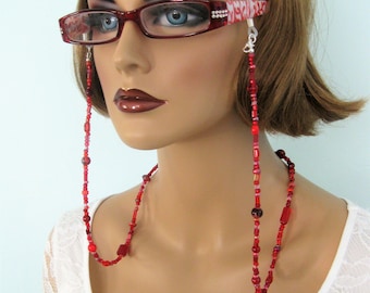 May include: Red beaded eyeglass chain with a silver clasp. The chain is made of small red beads and has a square-shaped bead at the end. The chain is worn by a person with red glasses.