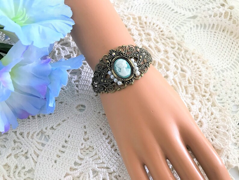 May include: A delicate bracelet featuring a cameo with a woman's profile in a light blue and white design. The bracelet is made of gold-toned metal and features a filigree design with white pearls.