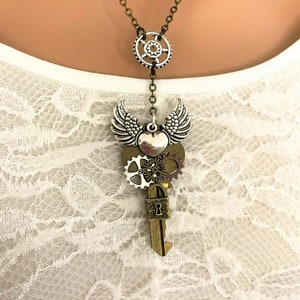 Peut inclure: Un collier argenté et bronze avec un pendentif en forme de clé, un cœur et des engrenages. La clé a un petit cadenas et le cœur a des ailes.