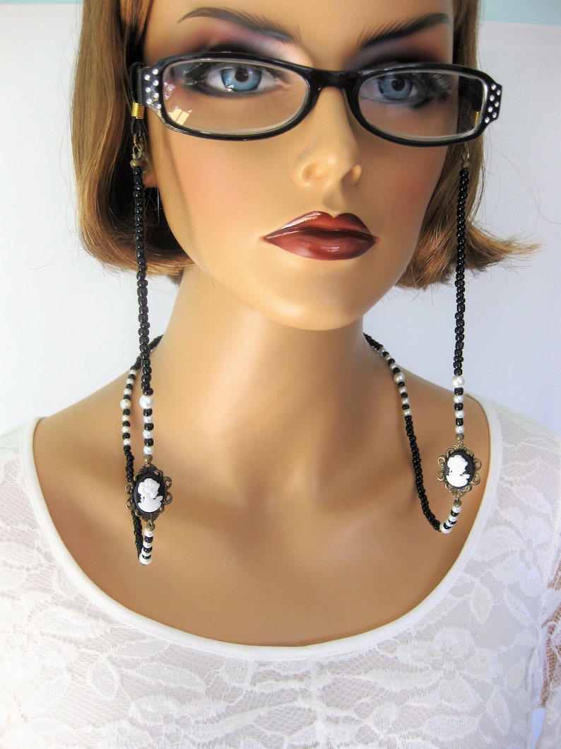 May include: Black and white beaded eyeglass chain with cameo charms. The chain is worn by a model with short brown hair and is attached to the temples of the glasses.